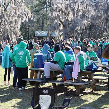 A large crowd of students gathers and socializes in an open area of a riverfront park